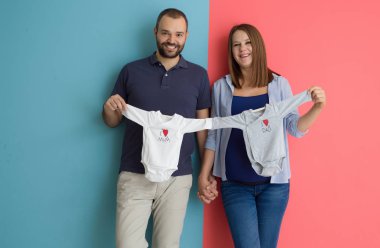 Güzel bir hamile kadın ve bebek bodysuits tutarak ve renkli arka plan üzerinde gülümseyen bekliyor bebek kocası