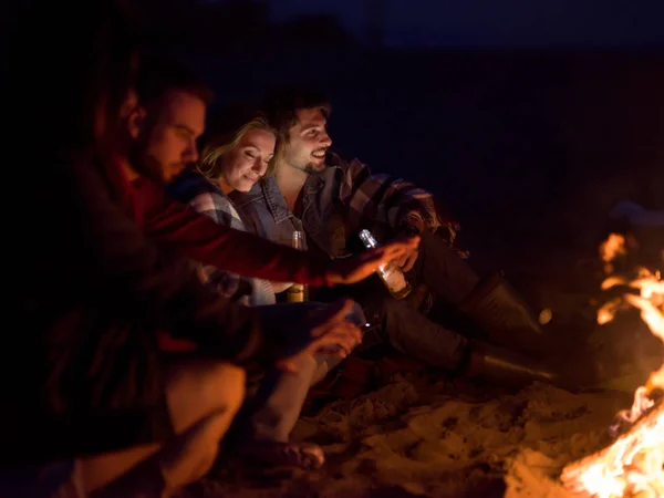 与朋友坐在海滩篝火边喝酒的年轻夫妇 — 图库照片