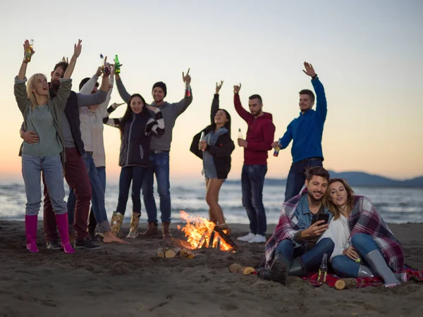 在秋天的海滩派对上 情侣们与朋友们一起喝啤酒玩乐 一起使用手机 — 图库照片
