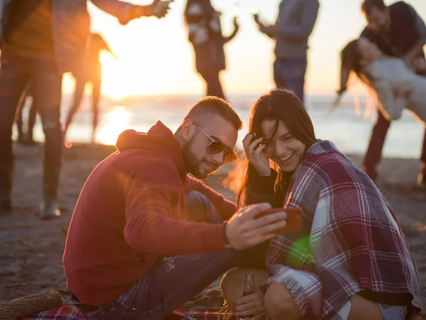 Pár Pomocí Mobilu Během Podzimní Plážové Párty Přáteli Popíjejícími Pivo — Stock fotografie