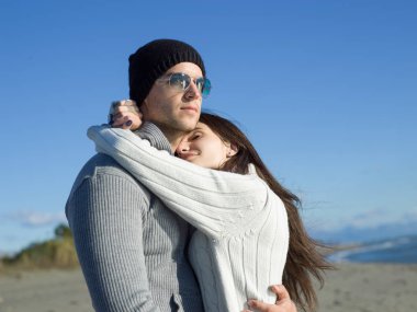 Genç çift, sonbahar güneşli bir günde kumsalda yürüyüp sarılıyor.