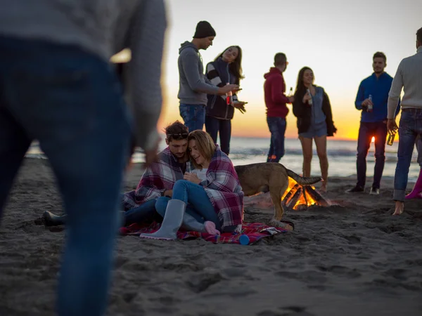 カップルビーチのキャンプファイヤーの周りの友人と一緒に座って日没時にビールを飲む — ストック写真