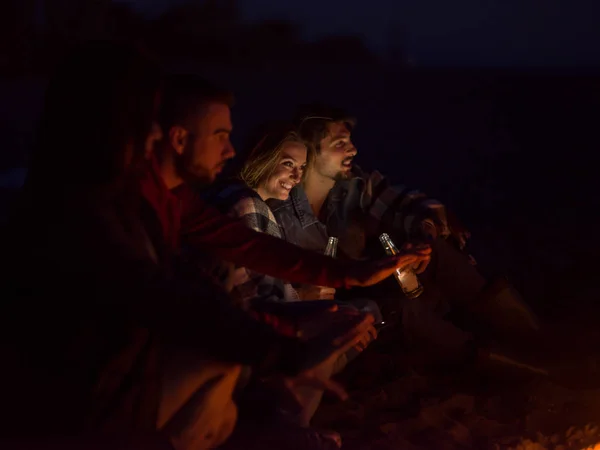 カップル夜のビーチでキャンプファイヤーの周りの友人と一緒に座ってビールを飲む — ストック写真