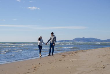 Genç çift, sonbahar güneşli bir günde kumsalda yürüyüp sarılıyor.