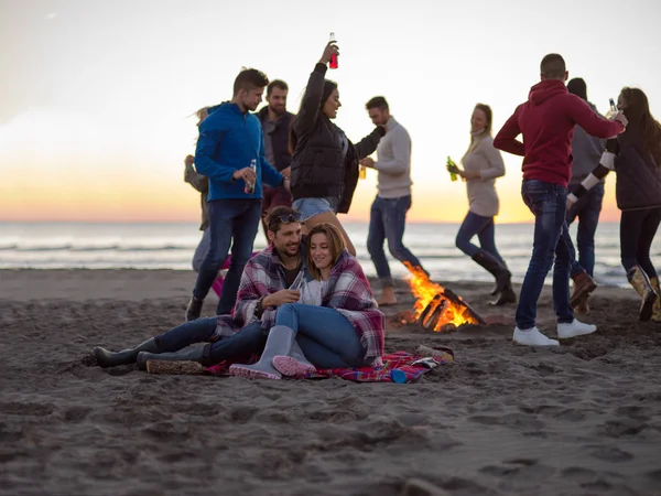 カップルビーチのキャンプファイヤーの周りの友人と一緒に座って日没時にビールを飲む — ストック写真