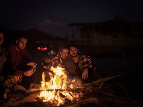与朋友坐在海滩篝火边的年轻夫妇晚上喝啤酒彩色滤水器 — 图库照片