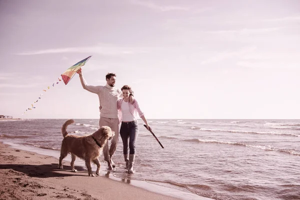 在秋天的日子里 一对年轻夫妇在海滩上与一只狗和一只风筝玩耍 玩得很开心 — 图库照片