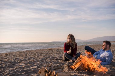 Genç çift ateşin yanında dinleniyor, sonbahar günü sahilde bira ya da içki içiyor.