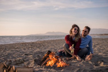 Genç çift ateşin yanında dinleniyor, sonbahar günü sahilde bira ya da içki içiyor.