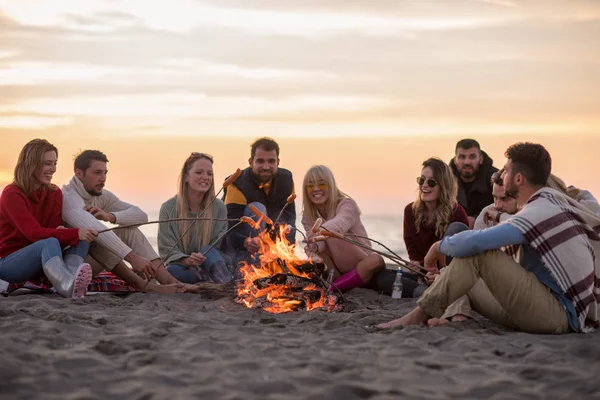 秋のビーチで火のそばに座ってソーセージを焼くとビールを飲む若い友人のグループは 話をし 楽しみを持って — ストック写真