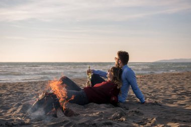 Genç çift ateşin yanında dinleniyor, sonbahar günü sahilde bira ya da içki içiyor.