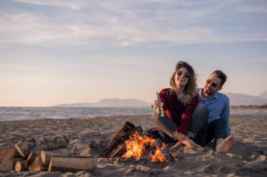Genç çift ateşin yanında dinleniyor, sonbahar günü sahilde bira ya da içki içiyor.