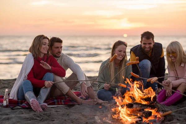 秋のビーチで火のそばに座ってソーセージを焼くとビールを飲む若い友人のグループは 話をし 楽しみを持って — ストック写真