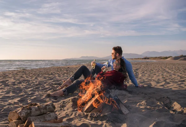 Young Couple Relaxing By The Fire, Drinking A Beer Or A Drink From The Bottle on the beach at autumn day