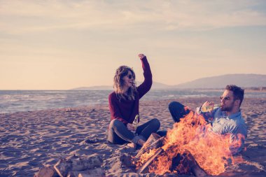 Genç çift ateşin yanında dinleniyor, sonbahar günü sahilde bira ya da içki içiyor.