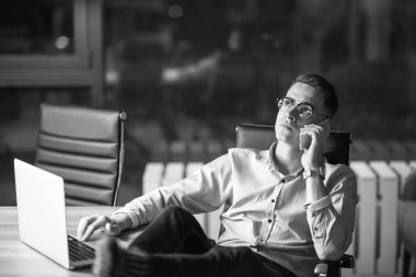 Genç işadamı geceleri karanlık ofiste dizüstü bilgisayarda çalışırken cep telefonu kullanıyor..