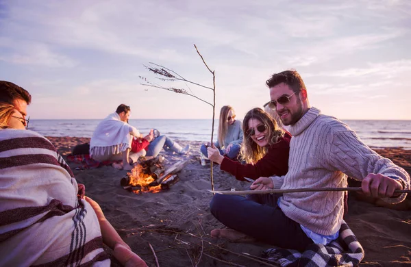 年轻的情侣和朋友们在海滩篝火边欢欢喜喜 日落时分喝啤酒滤水器 — 图库照片