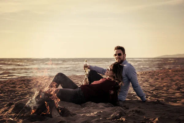 Young Couple Relaxing By The Fire, Drinking A Beer Or A Drink From The Bottle on the beach at autumn day filter