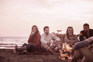 Bir grup genç arkadaş sonbahar kumsalında ateşin başında oturmuş sosis pişiriyor, bira içiyor, konuşuyor ve eğleniyorlar.