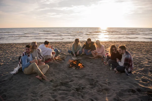 太陽が沈み始めるとビーチでたき火で楽しいビールを飲みながら幸せなケアフリー若い友人 — ストック写真