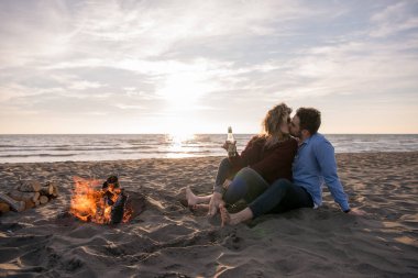 Genç çift ateşin yanında dinleniyor, sonbahar günü sahilde bira ya da içki içiyor.