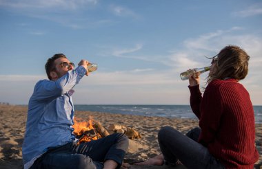 Genç çift ateşin yanında dinleniyor, sonbahar günü sahilde bira ya da içki içiyor.
