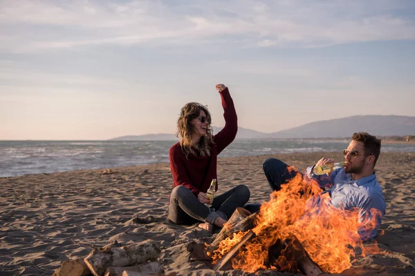 Young Couple Relaxing By The Fire, Drinking A Beer Or A Drink From The Bottle on the beach at autumn day