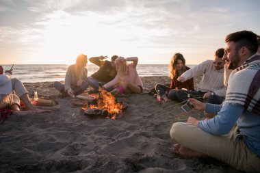 Mutlu tasasız genç arkadaşlar eğleniyor ve sahilde şenlik ateşiyle bira içiyor güneş batmaya başlarken