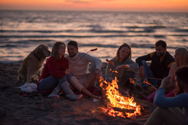 Group of young friends sitting by the fire at autumn beach, grilling sausages and drinking beer, talking and having fun
