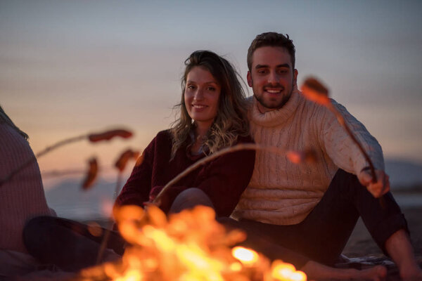 Group of young friends sitting by the fire at autumn beach, grilling sausages and drinking beer, talking and having fun