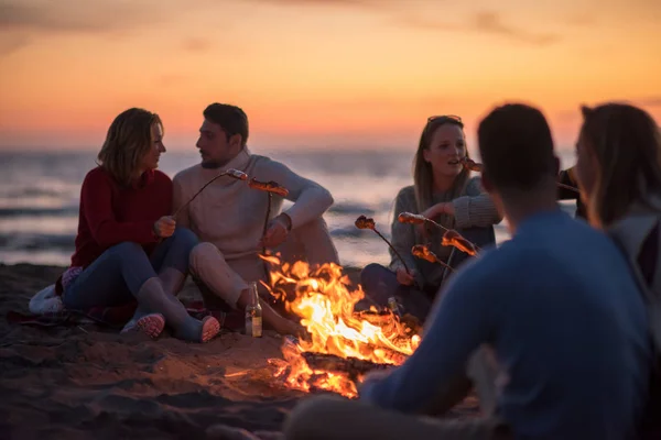 一群年轻的朋友坐在秋天海滩的篝火边 烤香肠 喝啤酒 — 图库照片