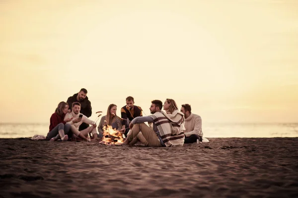 一群年轻的朋友坐在秋天海滩的篝火边 烤香肠 喝啤酒 玩乐地过滤 — 图库照片