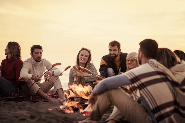 秋のビーチで火のそばに座っている若い友人のグループ ソーセージを焼くとビールを飲みます 話と楽しいフィルターを持っています — ストック写真