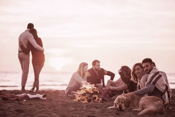 ビーチでキャンプファイヤーの周りの友人と楽しむ若いカップル日没時にビールフィルターを飲む — ストック写真
