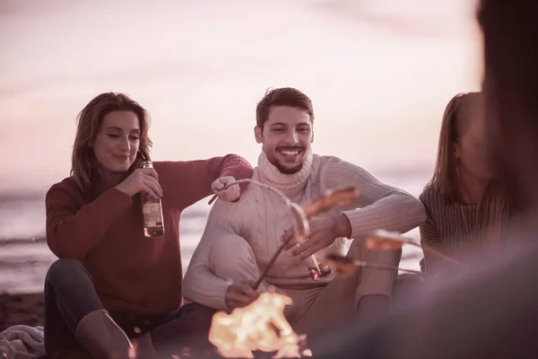 秋のビーチで火のそばに座っている若い友人のグループ ソーセージを焼くとビールを飲みます 話と楽しいフィルターを持っています — ストック写真