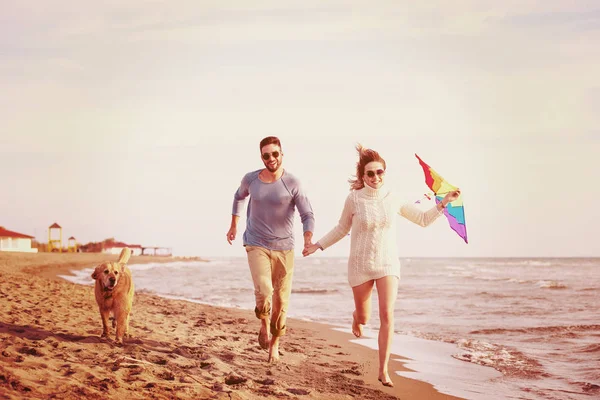 在秋天的日子里 一对年轻夫妇在海滩上与一只狗和一只风筝玩耍 玩得很开心 — 图库照片