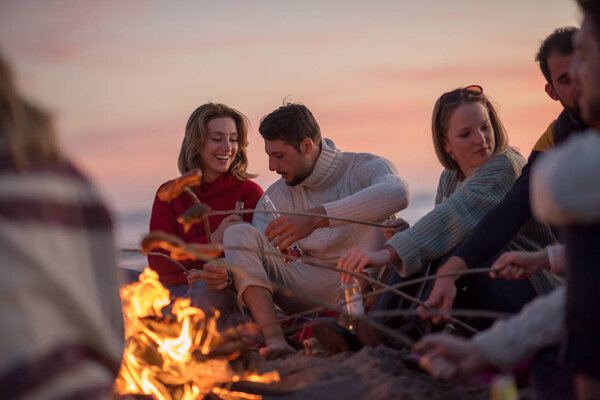 Group of young friends sitting by the fire at autumn beach, grilling sausages and drinking beer, talking and having fun