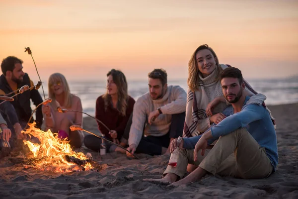 一群年轻的朋友坐在秋天海滩的篝火边 烤香肠 喝啤酒 — 图库照片