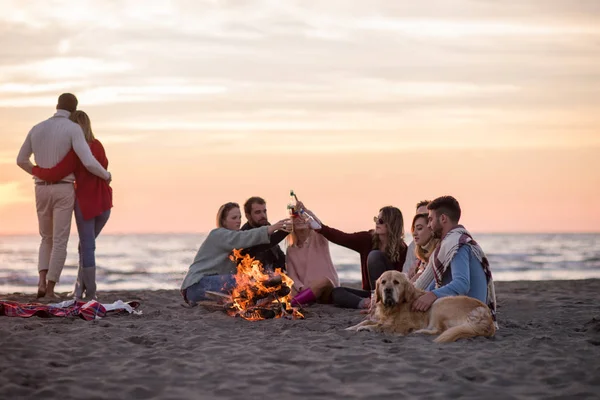 ビーチでキャンプファイヤーの周りの友人と楽しむ若いカップル日没時にビールを飲む — ストック写真