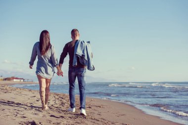 Genç çift, sonbahar güneşli bir günde kumsalda yürüyüp sarılıyor.