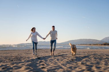 Plajda koşan çift, sonbahar gününde köpekle el ele tutuşuyor.