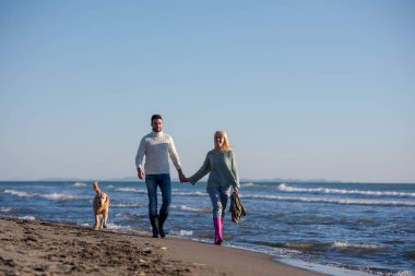 Plajda koşan çift, sonbahar gününde köpekle el ele tutuşuyor.