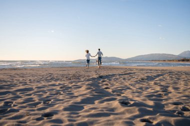 Plajda koşan çift, sonbahar gününde köpekle el ele tutuşuyor.