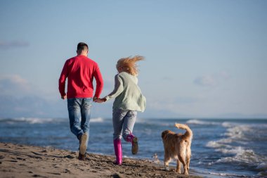 Plajda koşan çift, sonbahar gününde köpekle el ele tutuşuyor.