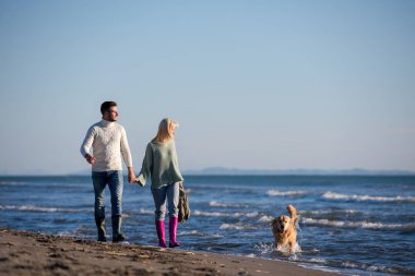 Plajda koşan çift, sonbahar gününde köpekle el ele tutuşuyor.