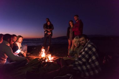 Genç çift gün batımında arkadaşlarıyla kumsaldaki kamp ateşinde bira içerek eğleniyor.