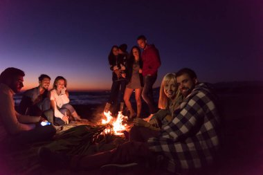 Genç çift gün batımında arkadaşlarıyla kumsaldaki kamp ateşinde bira içerek eğleniyor.