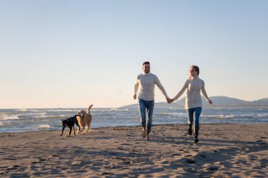 Plajda koşan çift, sonbahar gününde köpekle el ele tutuşuyor.