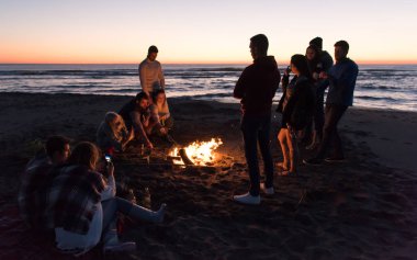 Mutlu tasasız genç arkadaşlar eğleniyor ve sahilde şenlik ateşiyle bira içiyor güneş batmaya başlarken