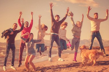 Bir grup heyecanlı genç arkadaş güneşli sonbahar kumsalında birlikte zıplıyorlar.